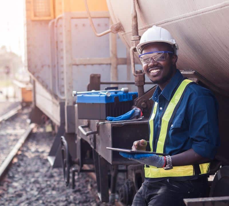 A man wearing a safety hat and vest leans against a tanker car