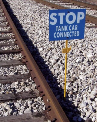The Spiked Hinged Staff is installed alongside a railroad track with a blue sign that reads: STOP TANK CAR CONNECTED