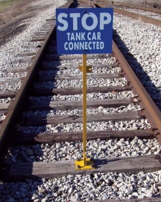 The Spiked Hinged Staff is installed on a railroad tie in the middle of the railroad track with a blue sign that reads: STOP TANK CAR CONNECTED
