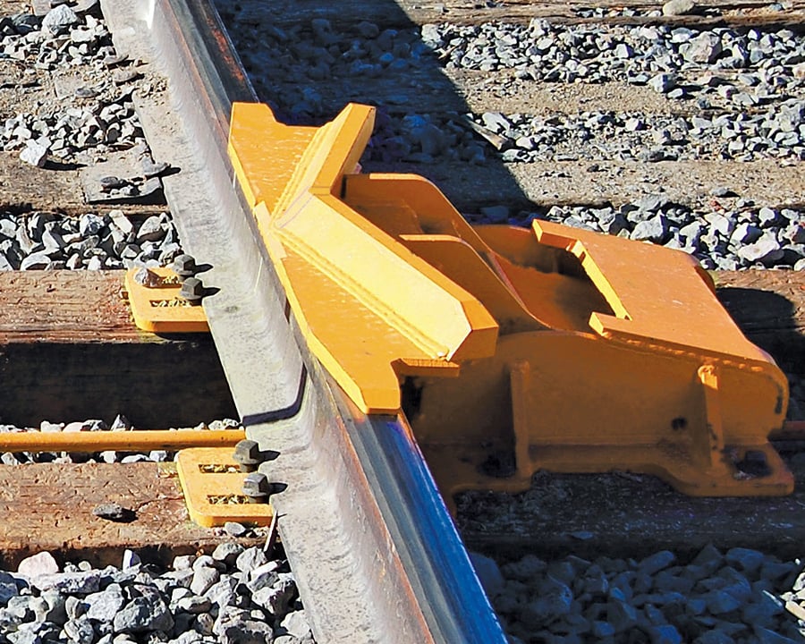 A large bright yellow, molded derailer clamped securely onto a steel rail set in a bed of gray gravel ballast.