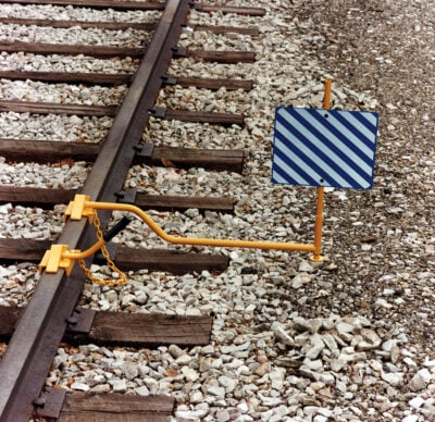 A yellow wheel chock assembly with a long handle and a blue-and-white diagonally striped sign is clamped to a railroad track.