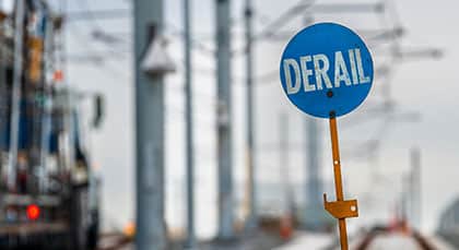 A circular blue sign reading "DERAIL" in white text stands on an orange post beside blurry train tracks.